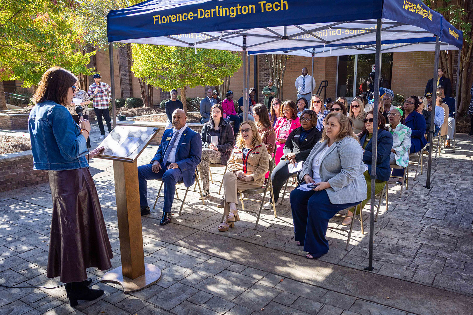 A group of people are seated in rows under a canopy while a speaker stands at a podium.